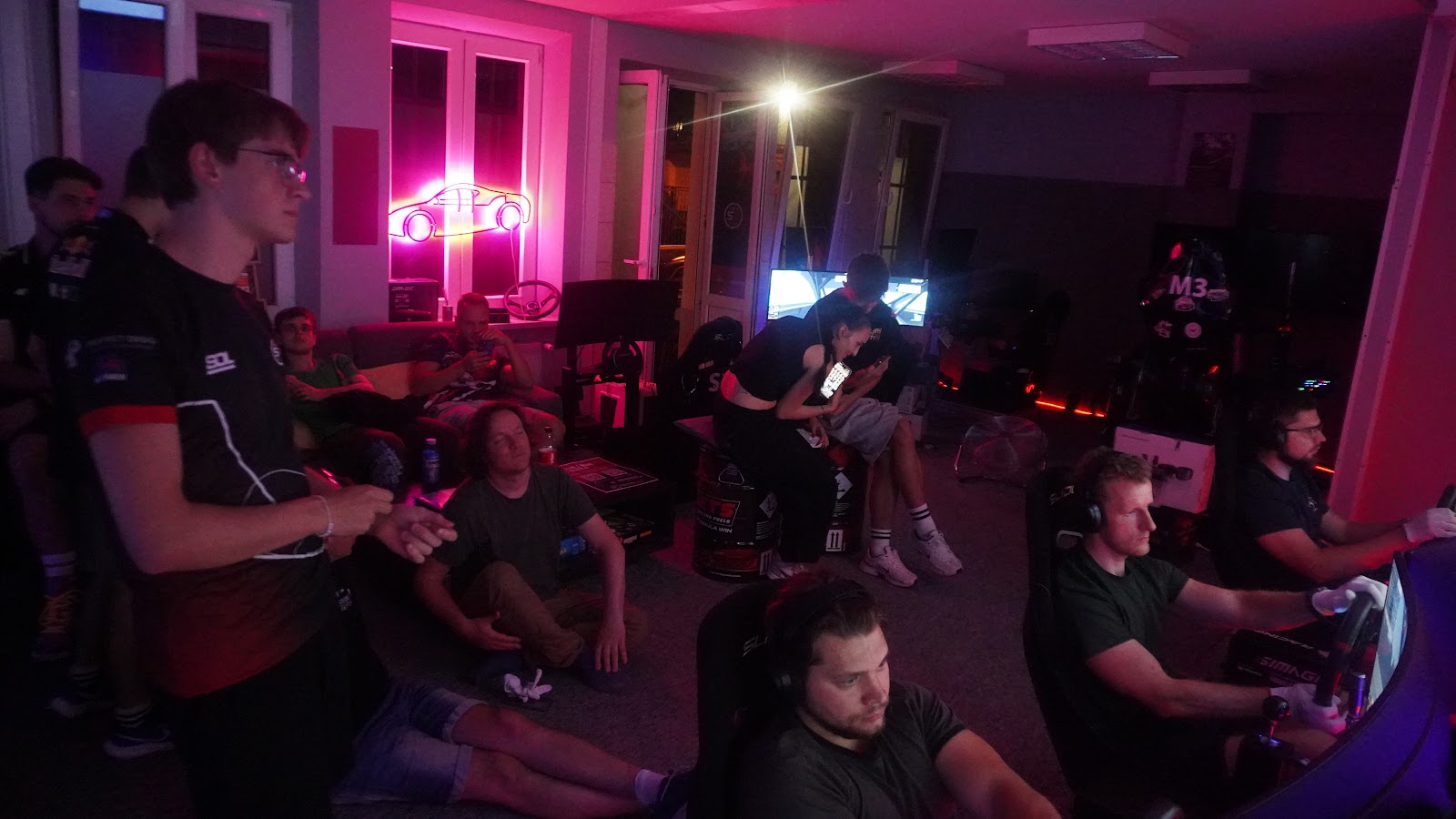 Szeroki widok sali SimRacing Dream z rzędem symulatorów zajętych przez kierowców ligowych w różowo-niebieskim świetle